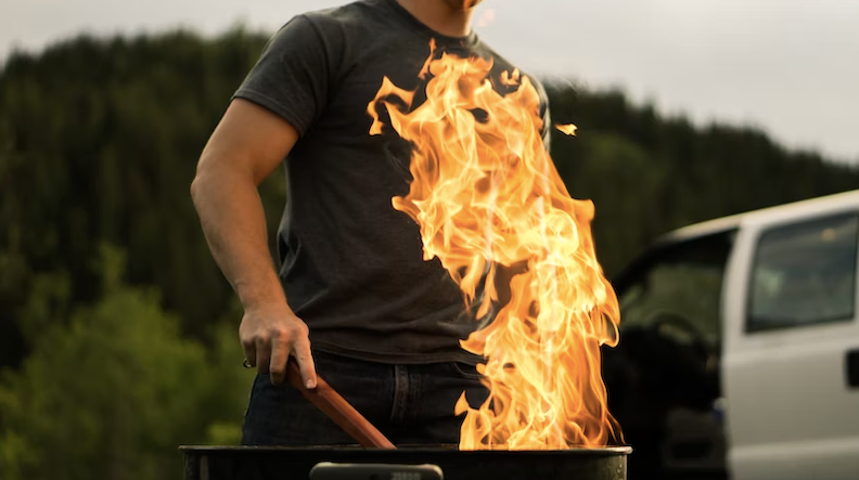 Man in dark t-shirt, cooking over the open flame of a grill.